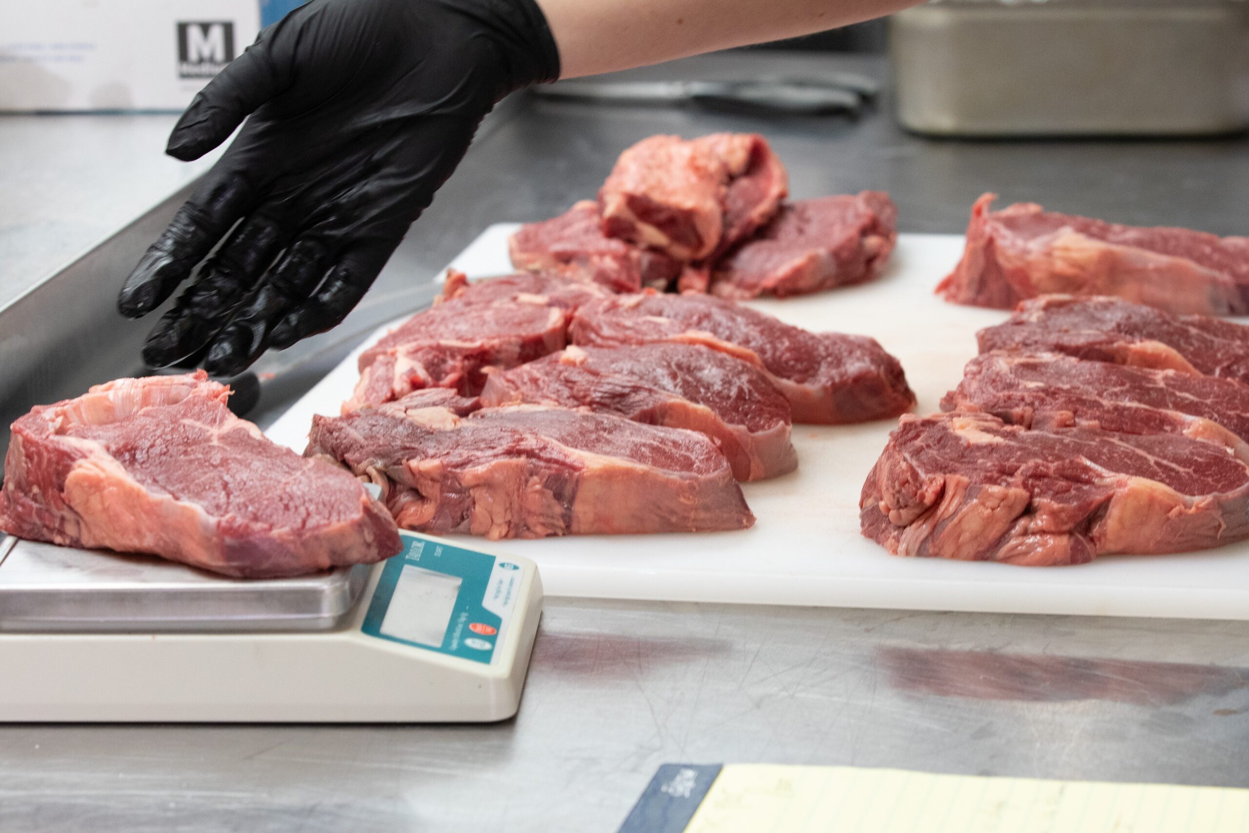 Several raw ribeye steaks are arranged on a white cutting board. A black-gloved hand hovers above one steak being weighed on a scale.