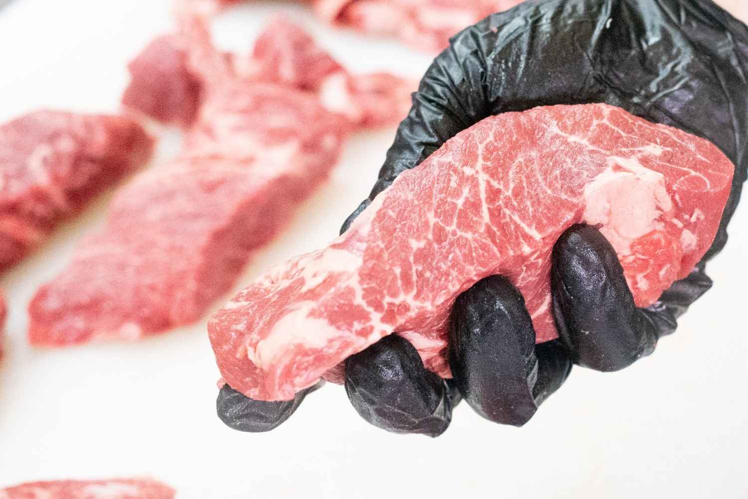 A black-gloved hand holds a raw Ocean Beef steak, displaying the white veins of fat that show its abundant marbling. Other Ocean Beef steaks sit on a white cutting board in the background 