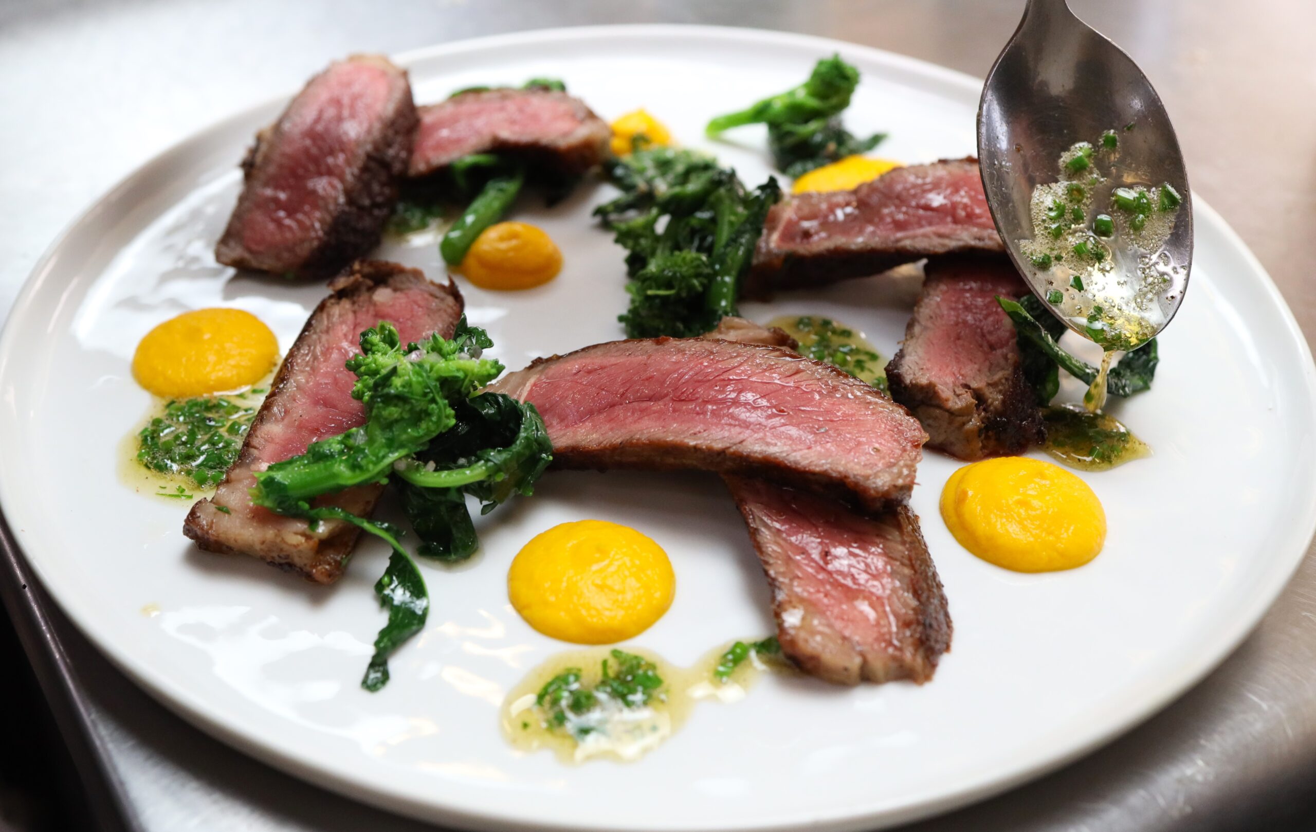 A spoon dribbles green herb sauce on a round white plate holding slices of cooked Ocean Beef steak with sauteed greens and dots of orange carrot puree.