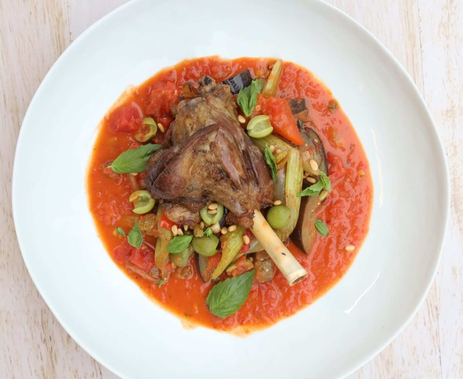 Braised Welsh Hill lamb foreshank on tomato puree with green olives and basil leaves in a white bowl on a light wood background.