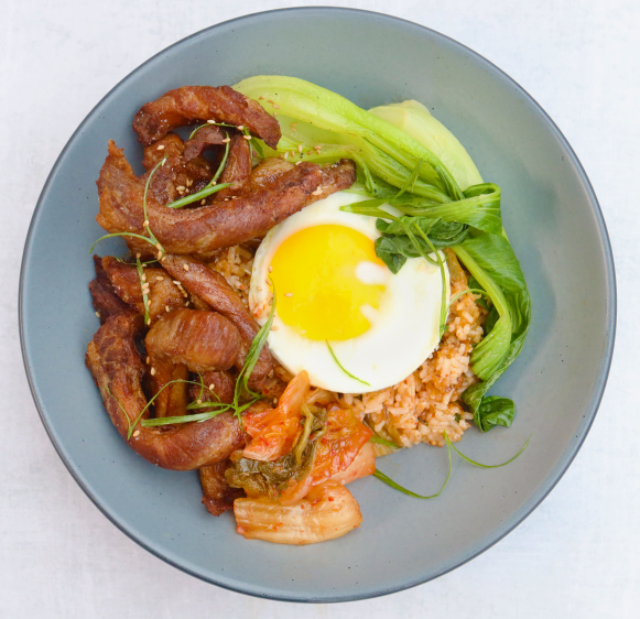 Iberico pork bibimbap with rice, kimchi, bok choy, and a fried egg in a blue bowl on a marble background.