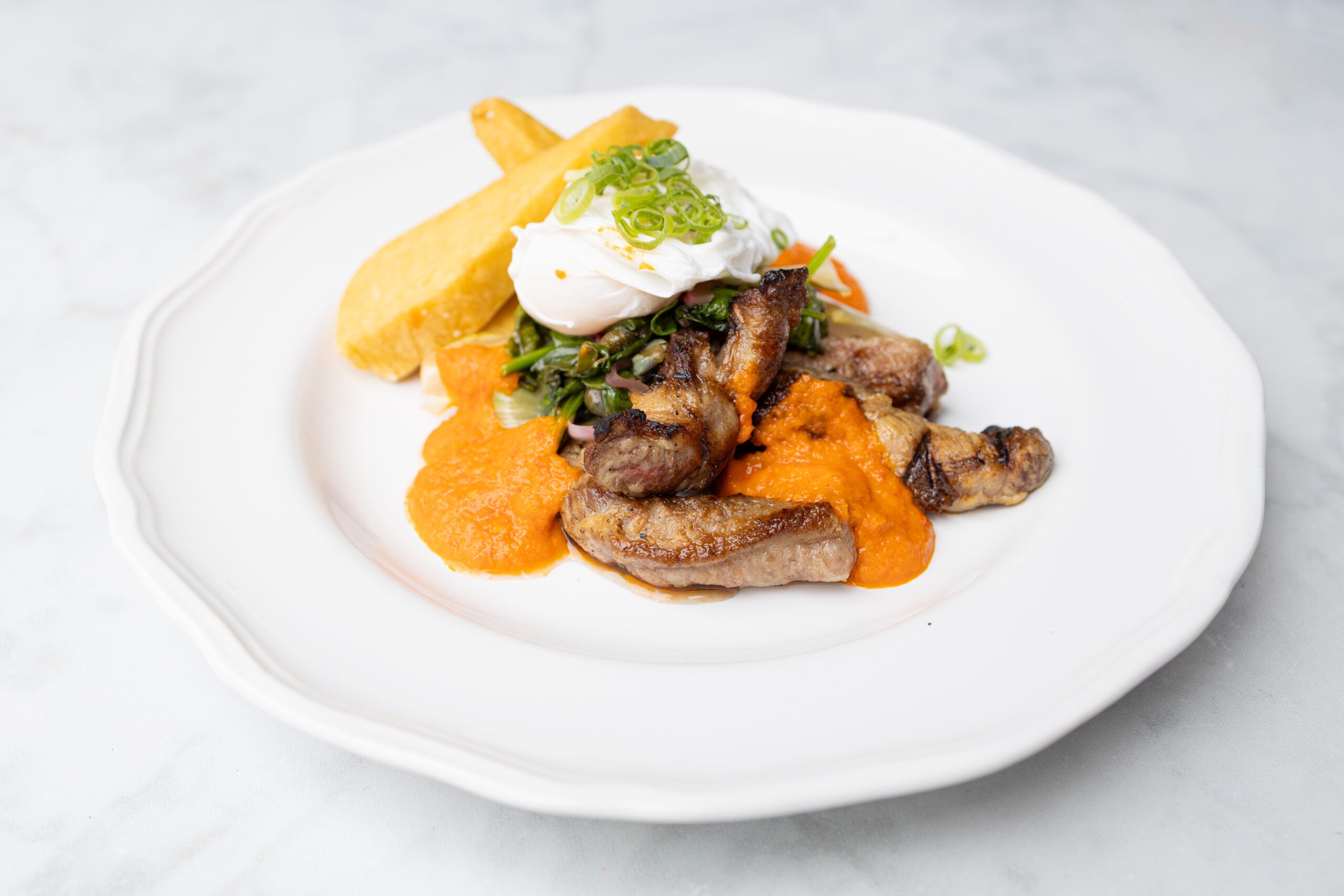 Grilled boneless Iberico pork strips with wilted greens, fried mashed chickpea wedges, orange harissa sauce, topped with a poached egg on a white plate.