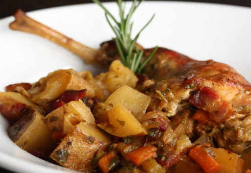 Braised Hermi rabbit leg with potatoes, carrots and a rosemary sprig in a white bowl on a black background.