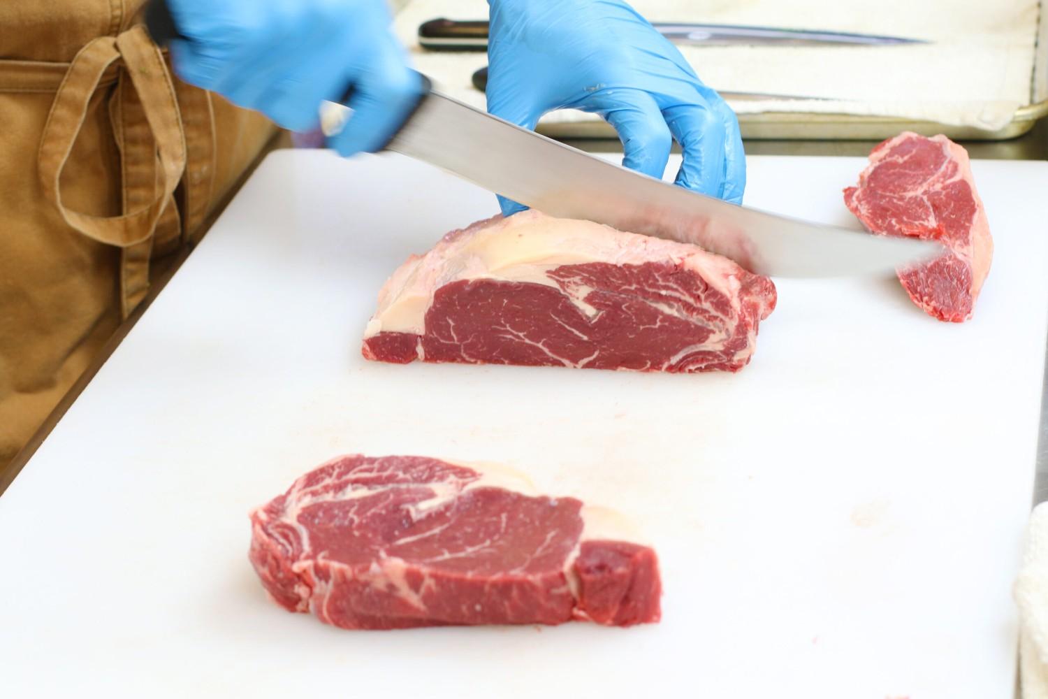 3.	A chef wearing a brown apron and blue gloves uses a curved knife to cut a raw Irish Nature Beef striploin into steaks on a white cutting board.
