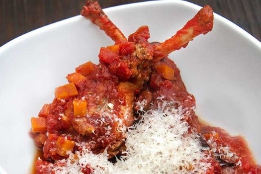 A square white bowl holds two crisscrossed rabbit legs smothered in a tomato sauce that is chunky with diced carrots and sprinkled with grated white cheese.