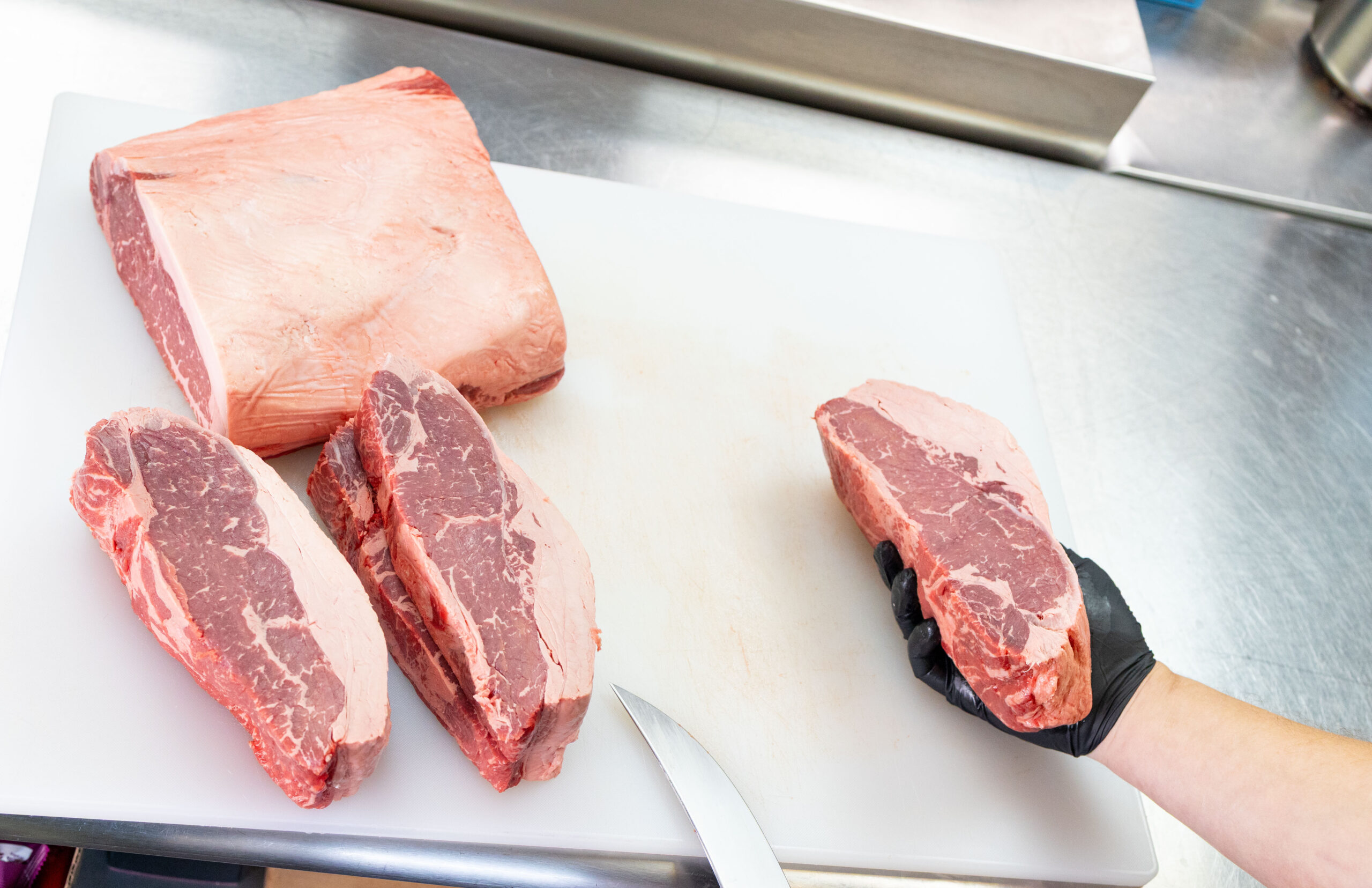 A black-gloved hand holds a raw strip steak. Two other strip steaks sit on a stainless-steel table, along with a larger, uncut portion of striploin subprimal. The blade of a knife appears in the foreground.