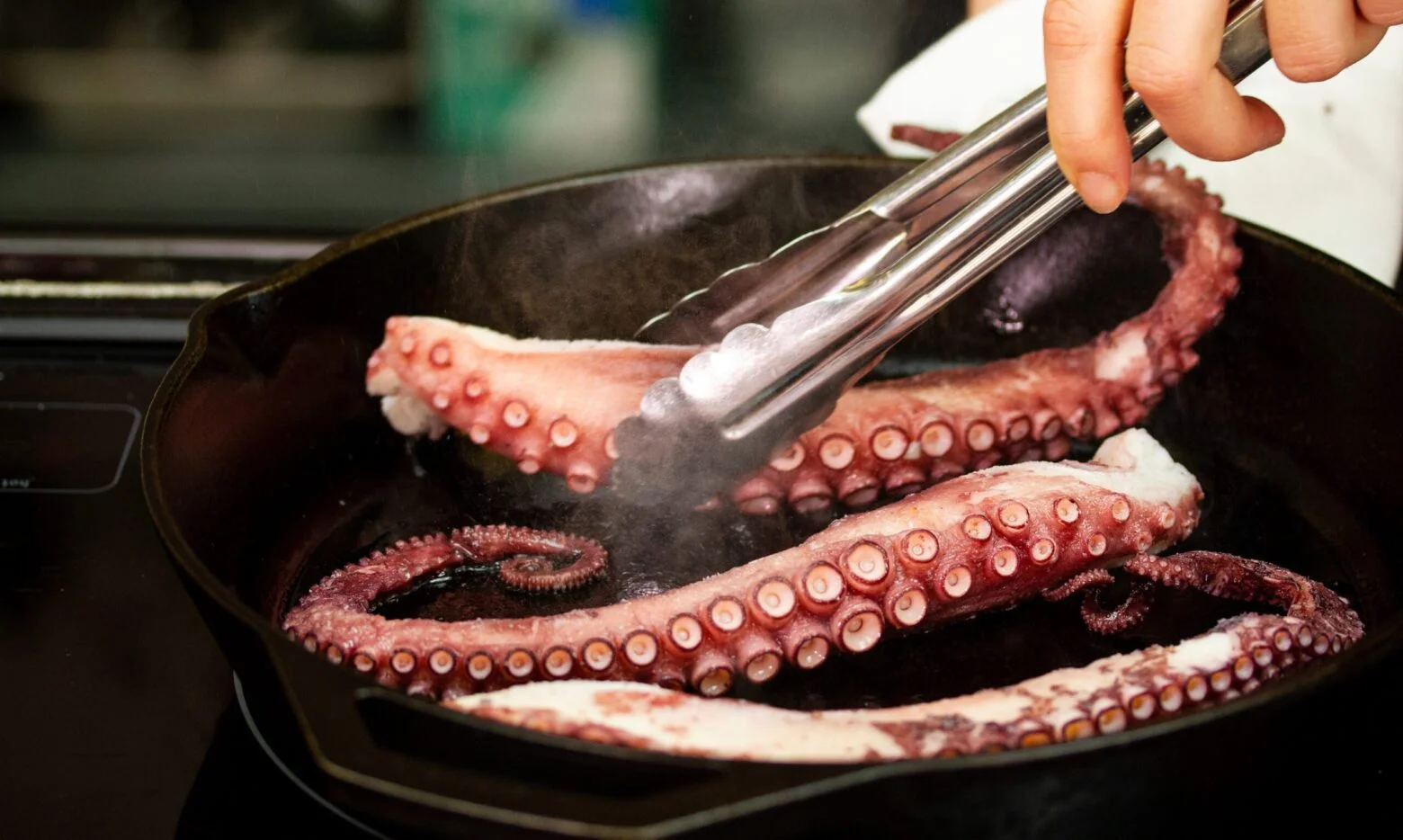 A close-up of octopus tentacles being cooked in a black cast-iron skillet. A hand is using metal tongs to turn the tentacles over. Steam rises from the pan, highlighting the vibrant pink and purple hues of the tentacles