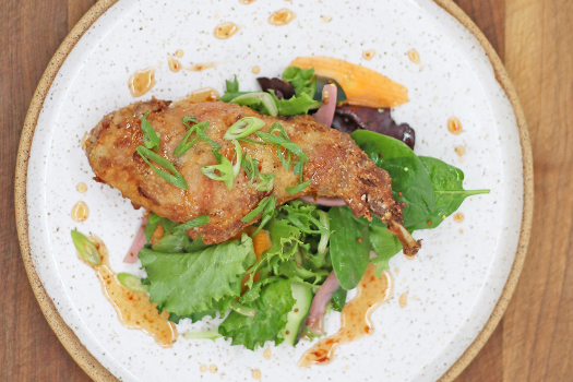 On a round white plate against a wood-grained backdrop, a chicken-fried rabbit leg sits atop salad greens drizzled with hot honey and an orange sauce.