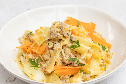 A white bowl holds pappardelle noodles tossed with rabbit Bolognese sauce, garnished with carrot curls and chopped parsley. 