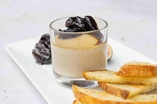 On a rectangular white plate, prunes stewed in Armagnac and a stack of toast points surround a glass container partially filled with rabbit liver mousse.