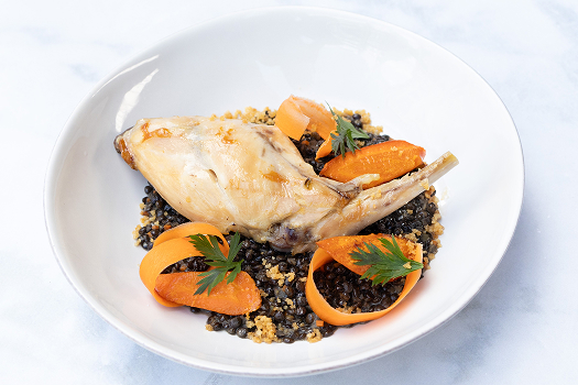 A white bowl holds a braised rabbit leg on a bed of black lentils with carrot curls and parsley sprigs. 