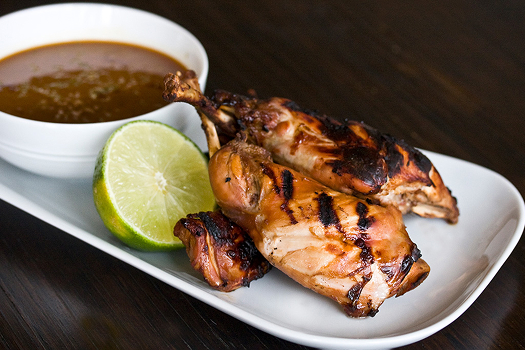 A rectangular white plate holds grilled rabbit pieces, half of a lime, and a bowl of brown Catalan stew. 