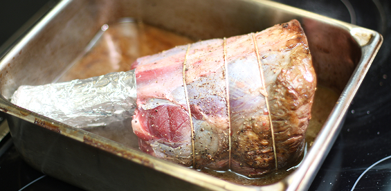 A seared Thor’s Hammer beef shank with aluminum foil wrapping its bone, sits on its side in a rectangular silver pan half full of braising liquid.