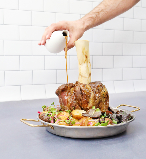 Against a background of white subway tiles, a cooked Thor’s Hammer beef shank sits upright in a round, two-handled pan surrounded by mixed vegetables as a hand pours sauce from a white pitcher. 