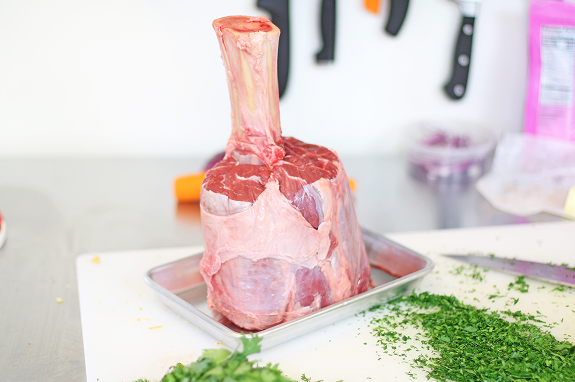 On a white kitchen counter, with knives hanging on the wall in the background and chopped green herbs in the foreground, a raw Thor’s Hammer beef shank sits upright on a rectangular silver sheet pan.