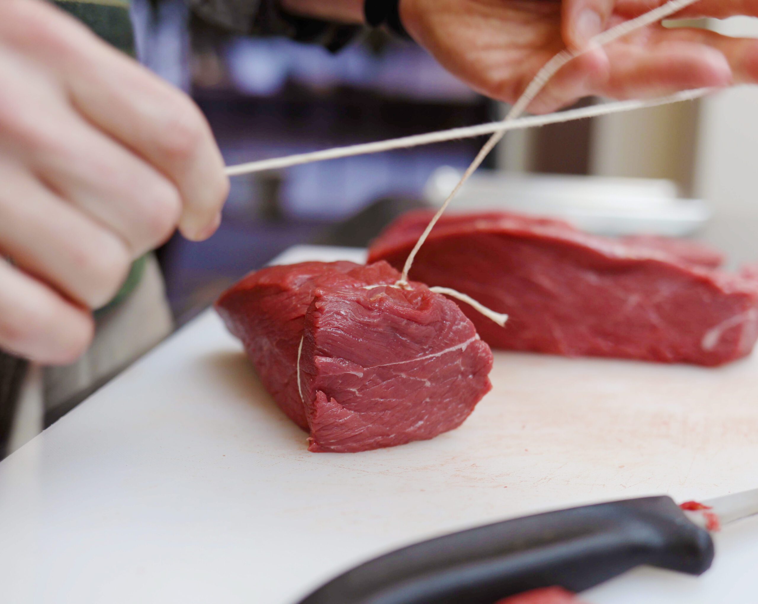 Two hands tying string around a Silver Fern Farms Angus beef filet on a white cutting board with a knife in the foreground.