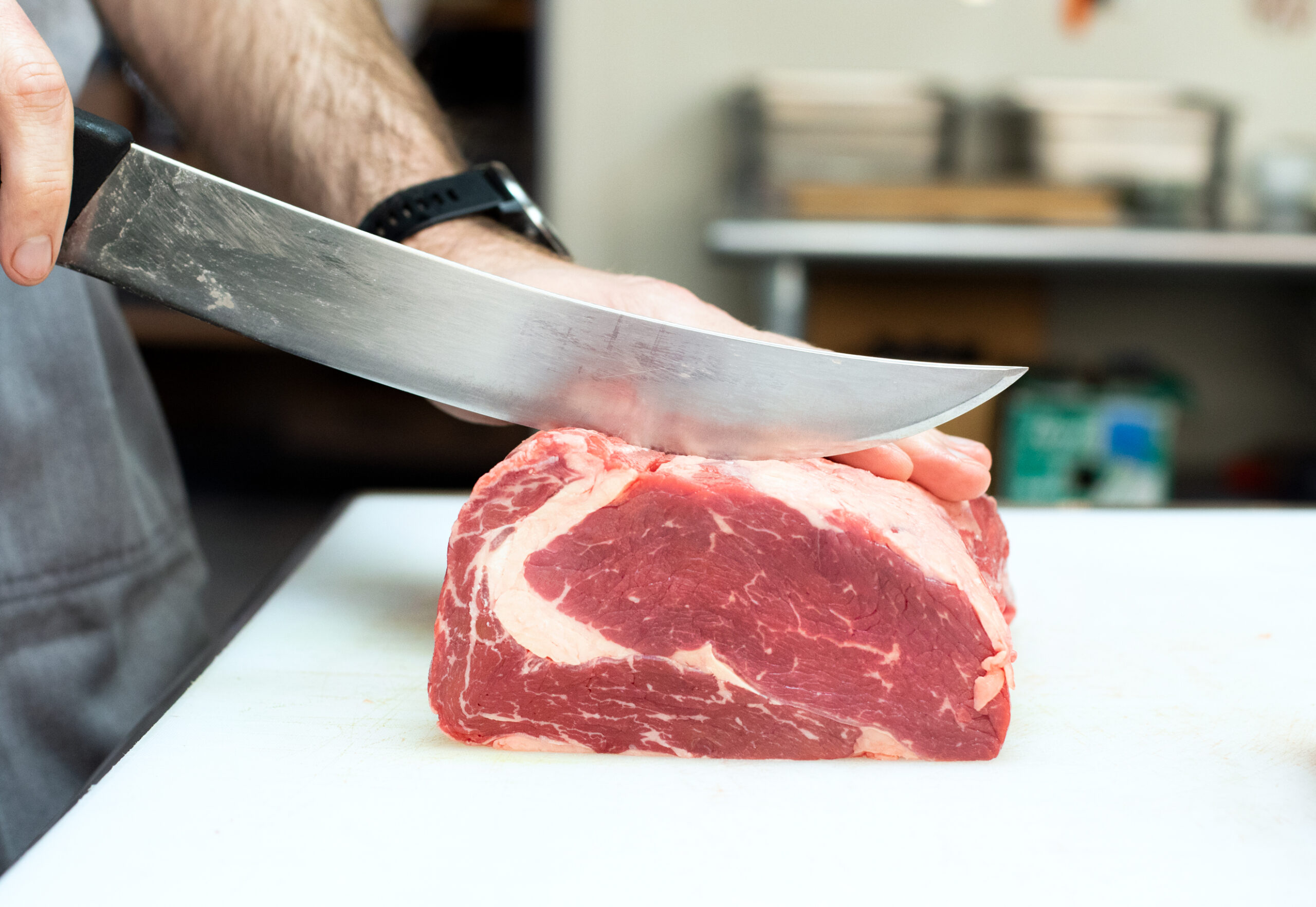 On a white cutting board, a curved knife blade cuts into a raw ribeye roast, held steady by a hand wearing a black wrist watch.