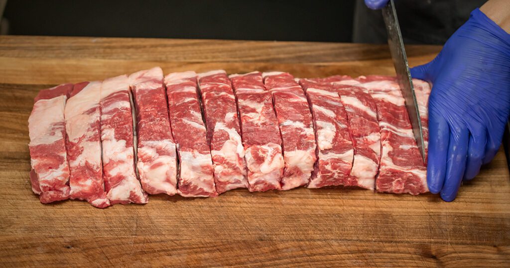 A blue gloved hand and a chef’s knife slicing a raw Silver Fern Farms whole ribeye roll into steaks.