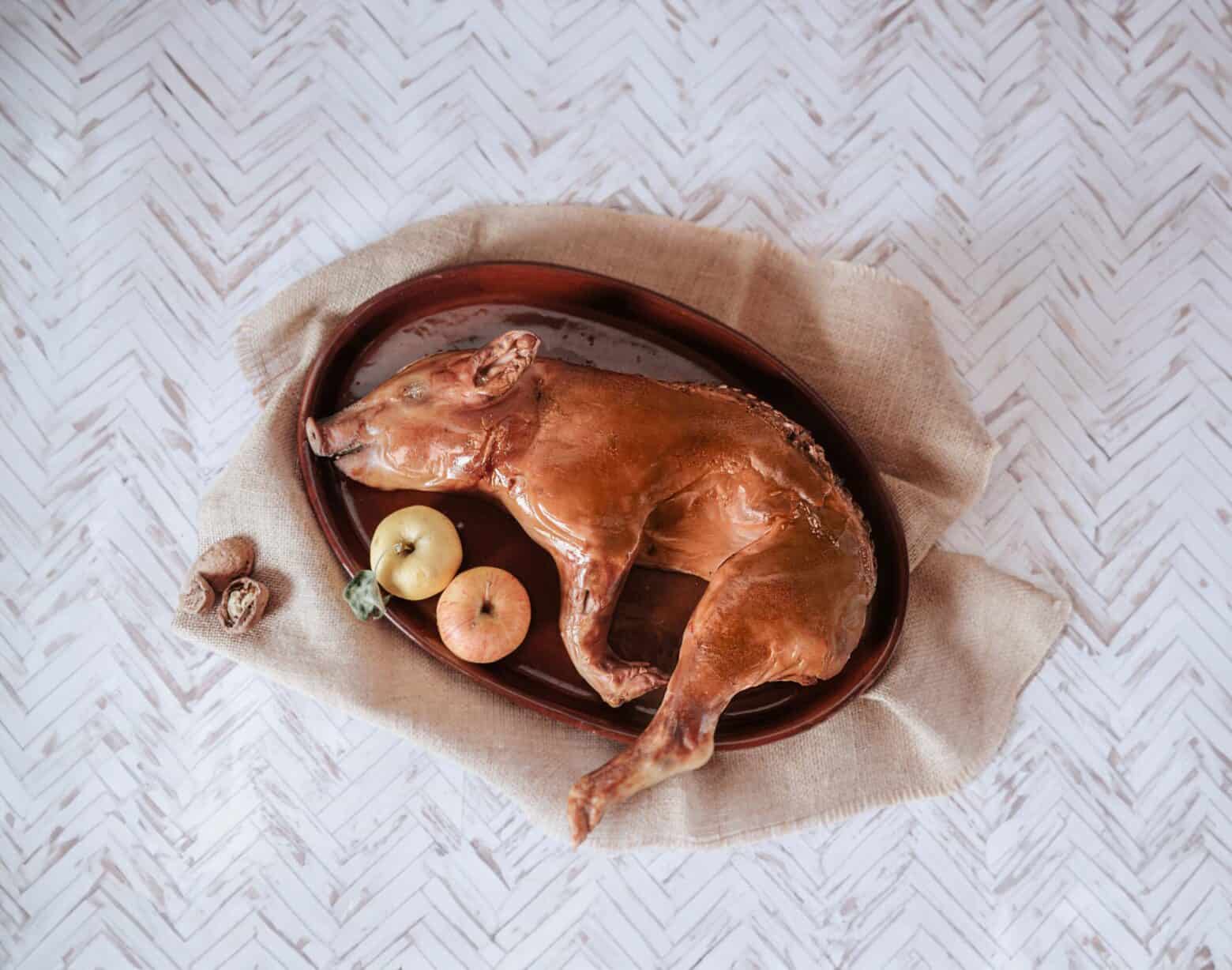 Against a white-washed chevron patterned backdrop, a whole roast suckling pig and 2 small apples are arranged on a brown ceramic oval platter set on a roughly textured beige cloth with three cracked walnuts in the lower left.