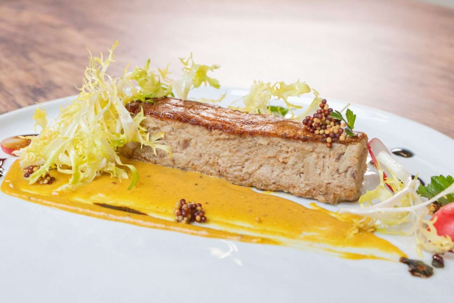 Slice of Cochinillo p&acirc;t&eacute; with mustard sauce, mustard seeds, and frisee on a white plate with a dark wooden background.