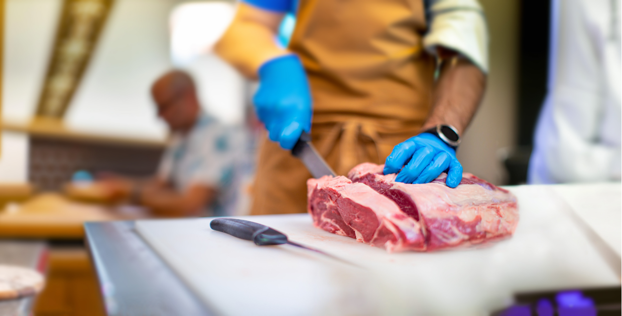 Two hands wearing blue gloves and a black watch on one arm slicing a raw Silver Fern Farms ribeye roll with a boning knife on a white cutting board.