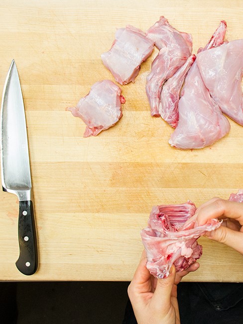 Two hands holding part of a butchered raw Hermi rabbit with other rabbit parts laying nearby on a wooden cutting board.