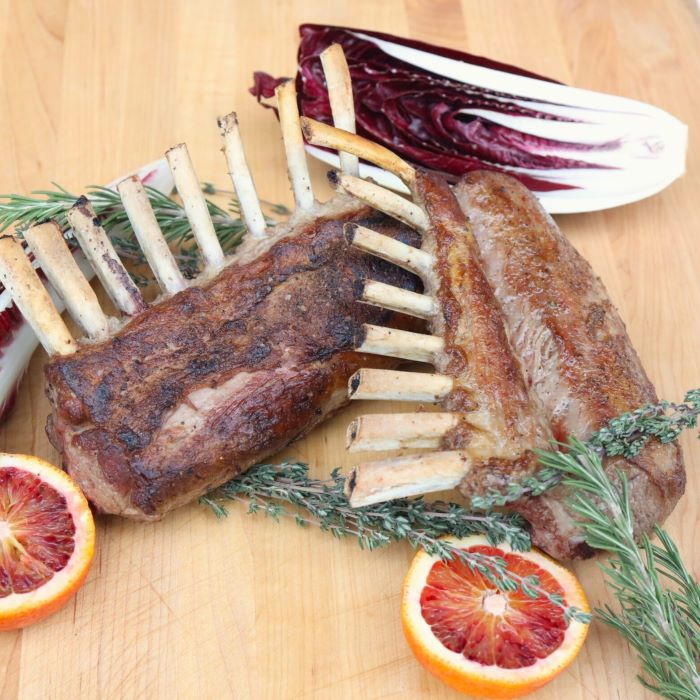 Two cooked Welsh Hill 8-bone lamb racks with half a blood orange, herb sprigs, and purple Treviso chicory on a wooden cutting board.