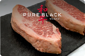 Two thick wagyu strip steaks on a black slate slab beneath the Pure Black Wagyu logo.