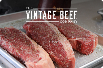 Three seasoned, mature-beef strip steaks on a silver pan beneath The Vintage Beef Company logo.