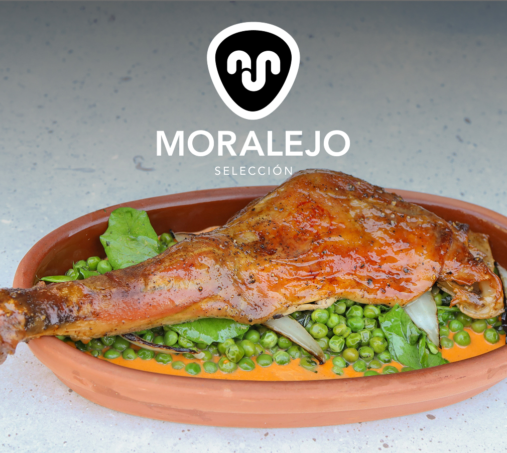 Beneath the Moralejo logo, an oval terra cotta dish holds a roasted suckling lamb leg with peas and onions and a roasted pepper sauce.