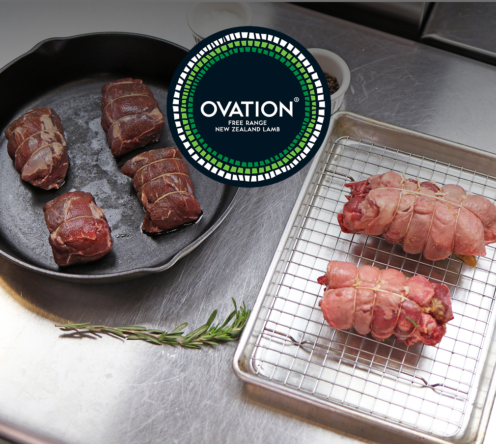 On a stainless-steel table, a cast iron skillet and a sheet pan hold boneless, raw Ovation lamb necks, stuffed and tied. A spring of fresh rosemary is at the bottom; the Ovation logo is at the top.