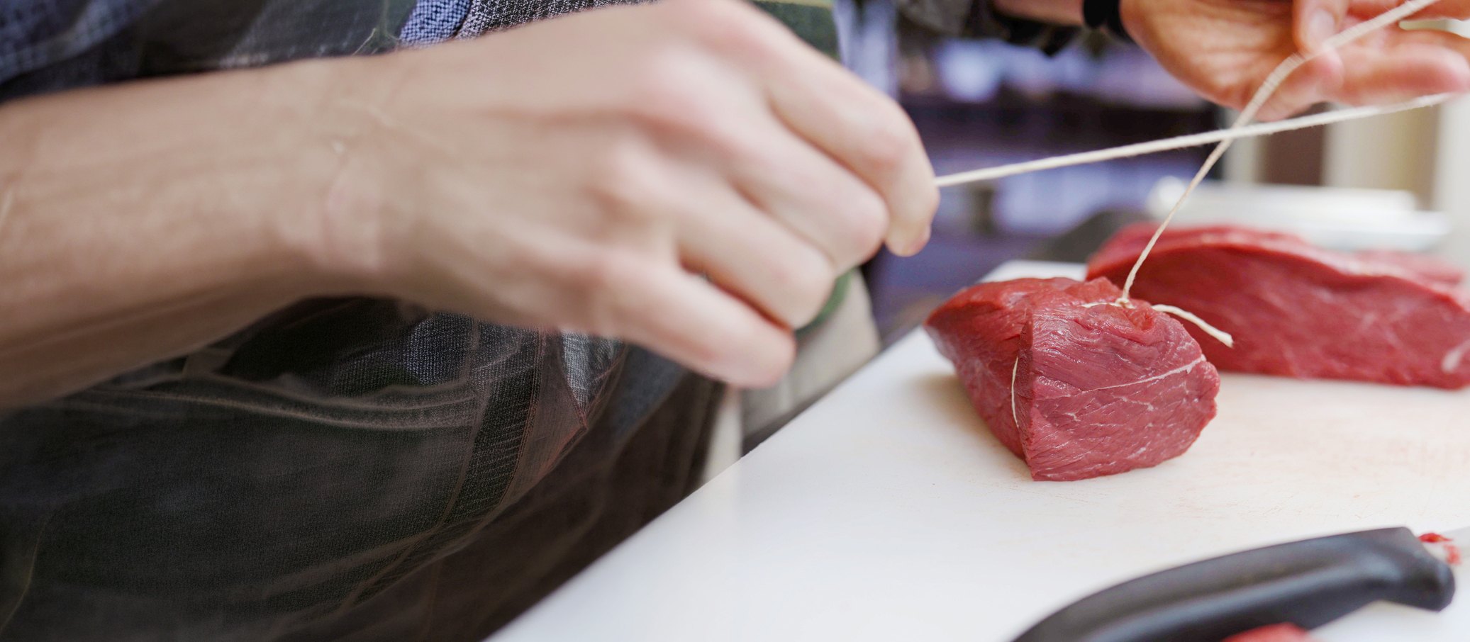 Two hands tying butcher&rsquo;s twine around a piece of raw meat.