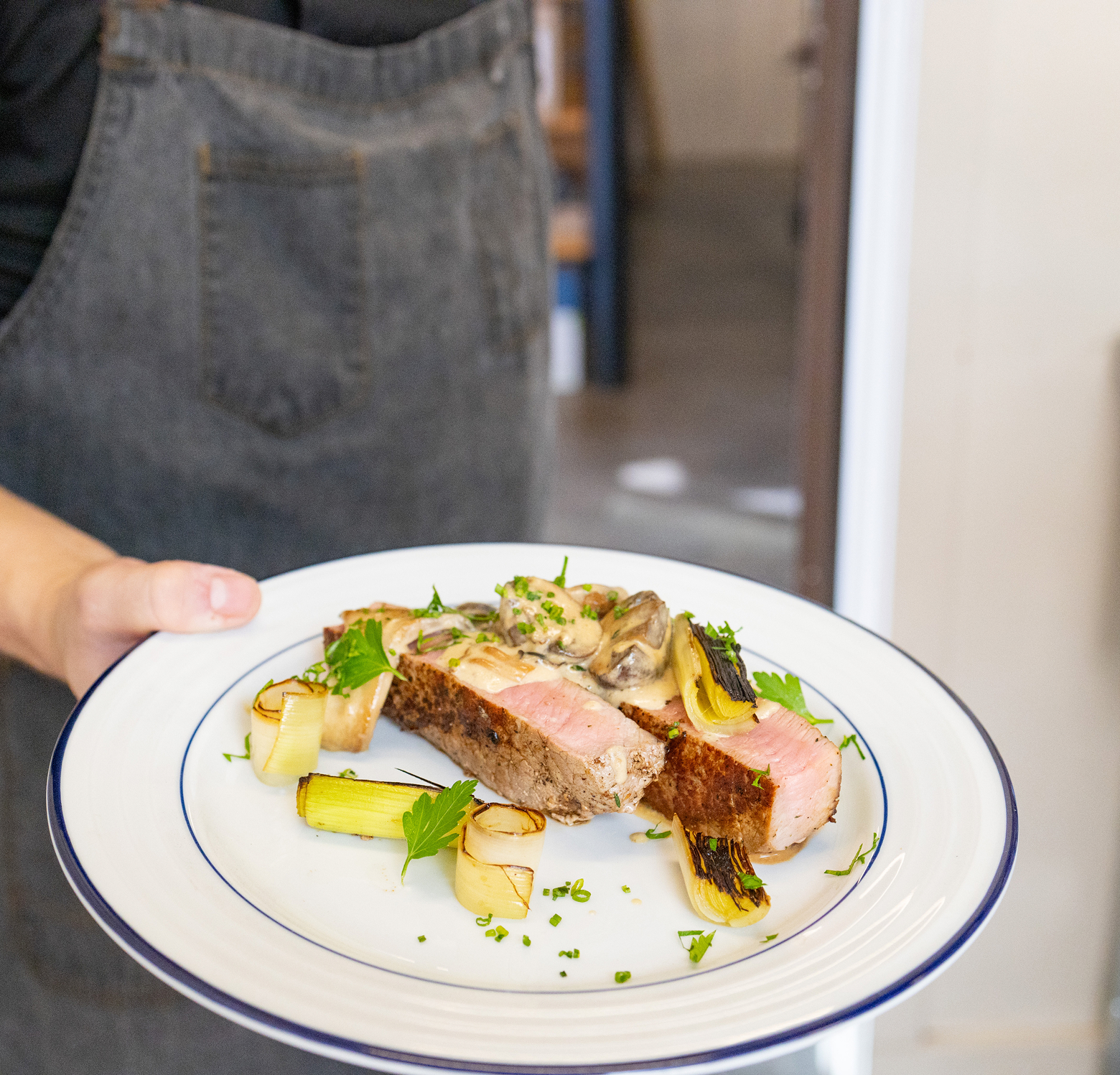 An aproned chef presents a plate of milk-fed veal with braised leeks and parsley on blue-rimmed white plate.
