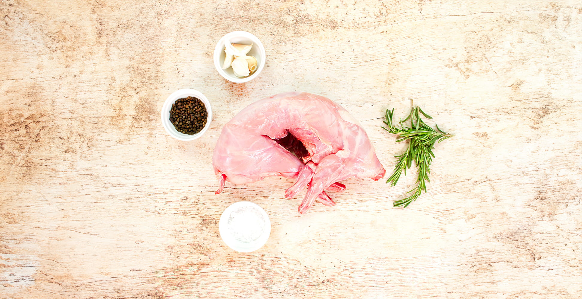 Rosemary sprigs and bowls of salt and peppercorns surround a whole, raw suckling pig lying curled on a whitewashed wooden board.